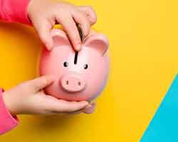 Child in pink sweatshirt putting coin into pink piggy bank over yellow and blue surface